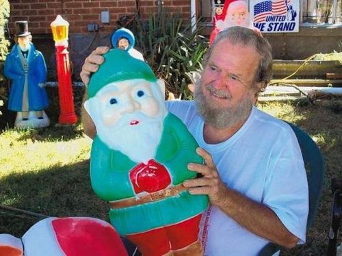 A man with a beard holds a colorful plastic Santa Claus figure in a yard decorated with holiday decorations.