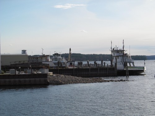 Two ferries docked at a pier, with calm water and a distant shoreline under a clear sky.