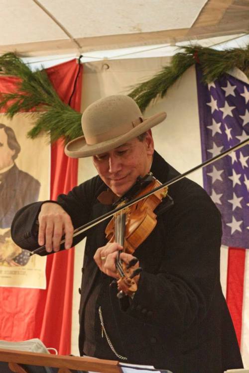 A musician in a hat plays the violin, performing in front of a festive backdrop with an American flag.