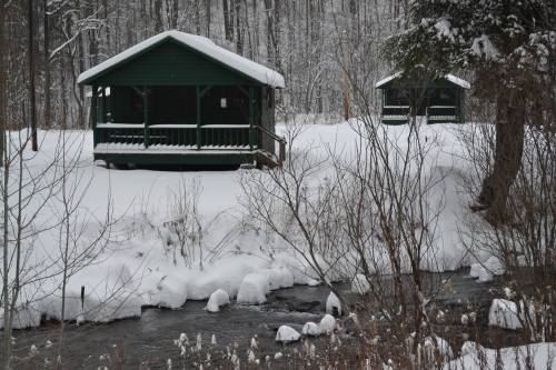 Two green cabins surrounded by snow, with a small stream flowing nearby in a winter landscape.