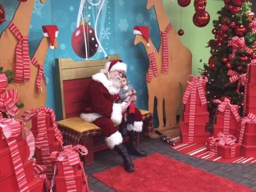 Santa Claus sits on a bench, holding a child, surrounded by festive decorations and wrapped gifts.