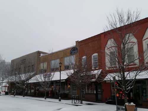 Snow-covered street with brick buildings and storefronts, trees bare, creating a quiet winter scene.