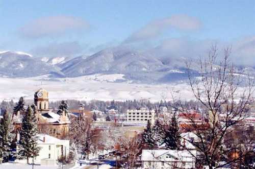 Snow-covered landscape with mountains in the background, featuring a clock tower and trees in the foreground.