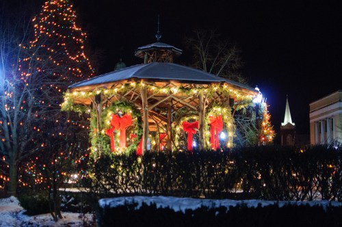A festive gazebo adorned with lights and greenery, surrounded by a decorated Christmas tree at night.