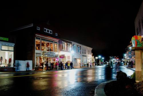 A festive street scene at night, with shops decorated in lights and a wet road reflecting the colorful displays.