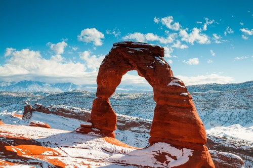 A snow-dusted red rock arch stands against a blue sky with clouds, surrounded by a winter landscape.