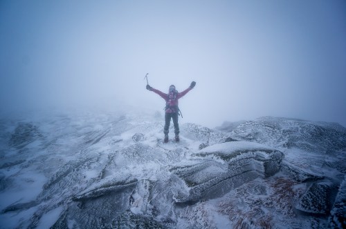 A climber stands triumphantly on a snowy, rocky peak, surrounded by fog and mist.