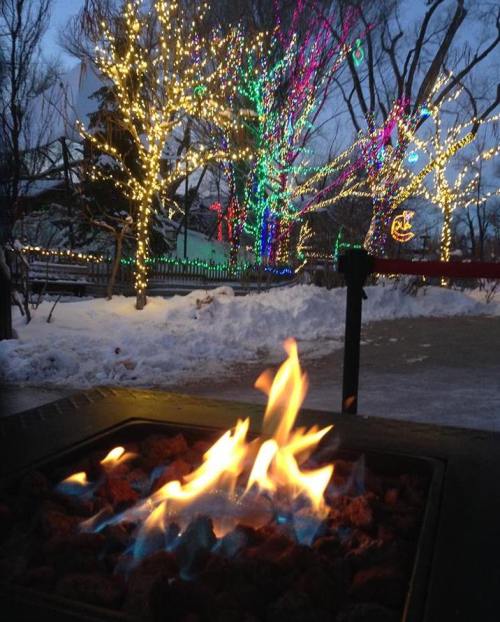 A cozy fire pit with blue and orange flames, surrounded by snow and colorful holiday lights on trees in the background.