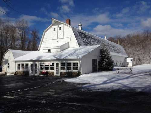 A large white barn covered in snow, surrounded by trees and a clear blue sky.