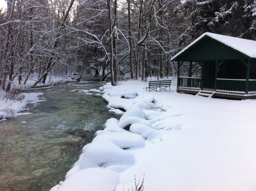 A snowy landscape featuring a green cabin by a flowing river, surrounded by trees and a blanket of fresh snow.
