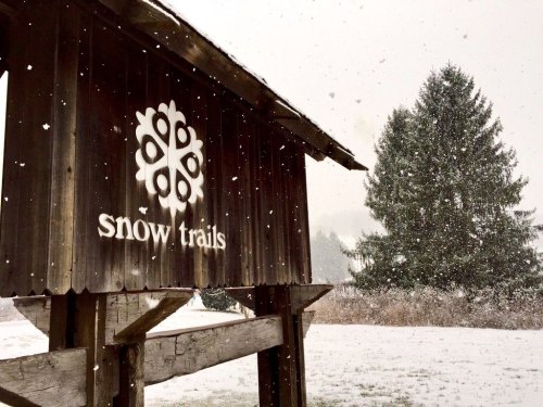 A wooden sign reading "snow trails" amidst falling snow, with a snowy landscape and evergreen trees in the background.
