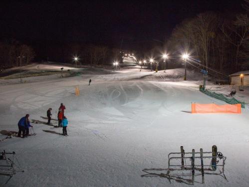 Skiers on a snow-covered slope at night, illuminated by bright lights, with a clear path and trees in the background.