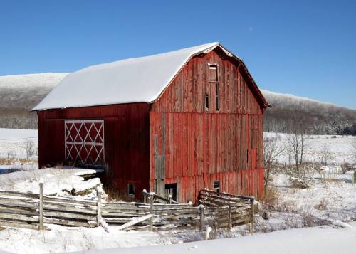 A red barn covered in snow, surrounded by a snowy landscape and a wooden fence under a clear blue sky.