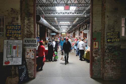 A bustling indoor market with people browsing tables, surrounded by brick walls and signs for local art and services.