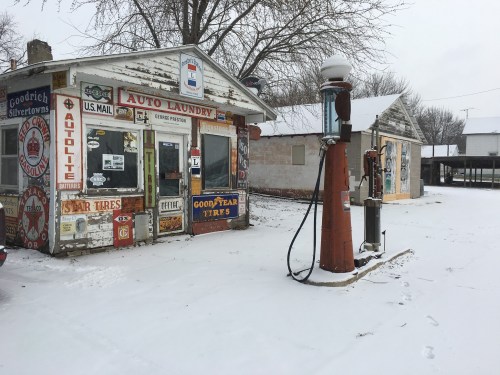 A vintage auto laundry with colorful signage and an old gas pump, surrounded by snow-covered ground and bare trees.