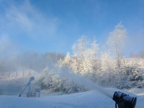 A snow-making machine sprays artificial snow onto a snowy landscape with frosted trees under a clear blue sky.