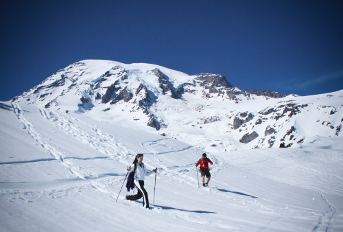 Two people hike on a snowy mountain trail under a clear blue sky, with a large peak in the background.