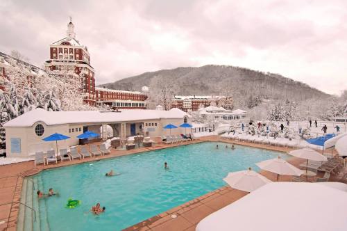 A snowy landscape surrounds a pool area with people swimming, umbrellas, and a historic building in the background.