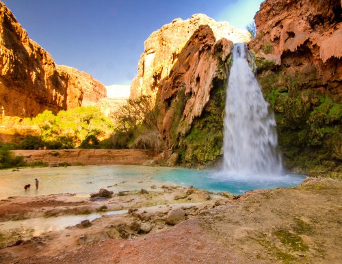 A stunning waterfall cascades into a turquoise pool, surrounded by red rock cliffs and lush greenery.