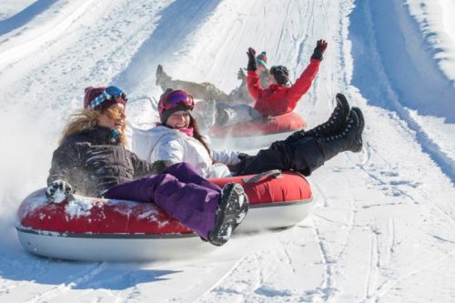 Three people enjoy snow tubing down a snowy slope, laughing and having fun in winter gear.