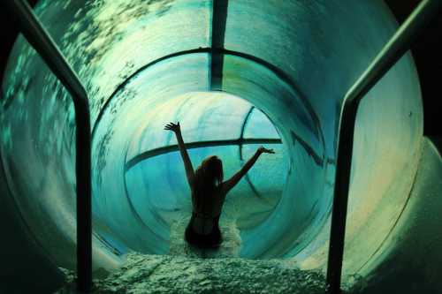 A person with arms raised slides down a water slide, surrounded by blue-green water and light.