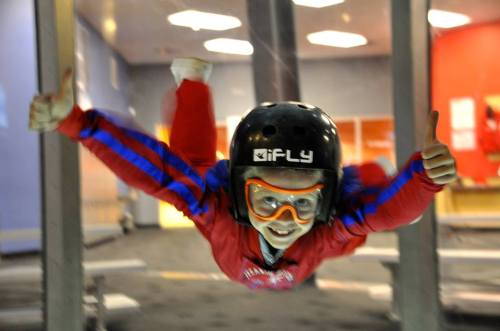 A child in a helmet and goggles smiles while flying in a wind tunnel, giving a thumbs-up.