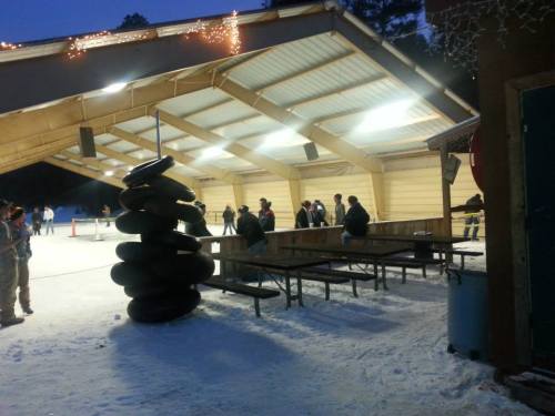 A snowy outdoor ice skating rink with a covered area, people gathered, and stacked inner tubes nearby.
