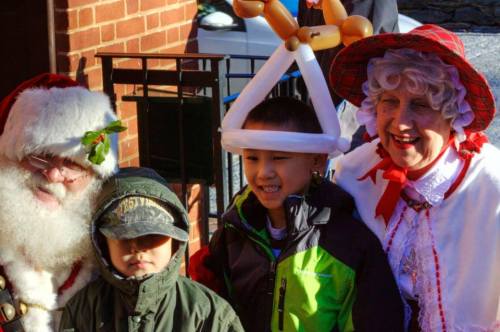 Santa Claus and a woman in festive attire pose with two children, one wearing a balloon hat, outside a brick building.