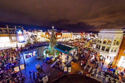 A festive night scene of a crowded town square decorated for Christmas, featuring a large tree and holiday lights.