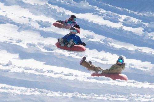 Three people sledding down a snowy hill on red inflatable tubes, surrounded by white snow and a clear blue sky.