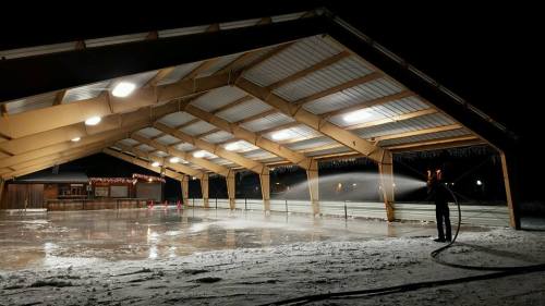 A person sprays water on an outdoor ice rink under a large covered structure at night.