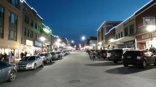 A bustling street at dusk, lined with shops and parked cars, illuminated by streetlights and festive lights.