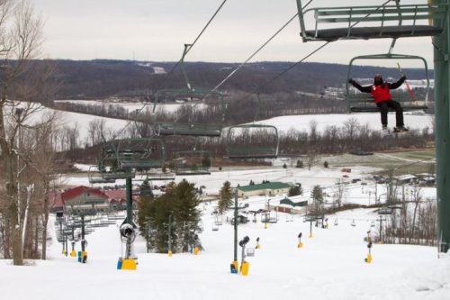 A skier on a chairlift overlooking a snowy slope and trees, with buildings in the background.