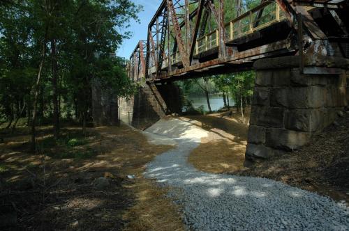 An old, rusted bridge spans a river, surrounded by trees and a gravel path leading to the water's edge.