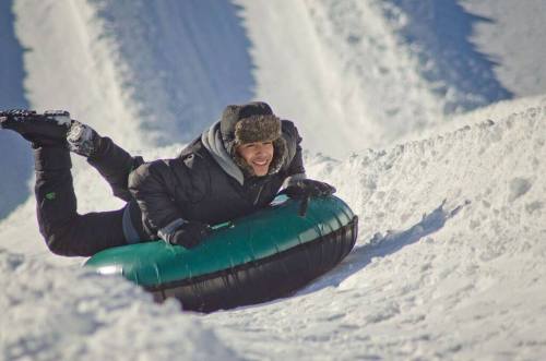 A person in winter gear lies on a green snow tube, sliding down a snowy slope.