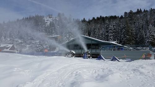 Snowmaking machines spray artificial snow near a ski lodge, surrounded by a snowy landscape and pine trees.