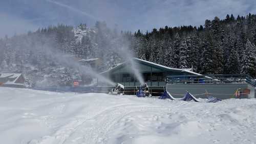 Snowmaking machines spray artificial snow near a ski lodge, surrounded by a snowy landscape and pine trees.