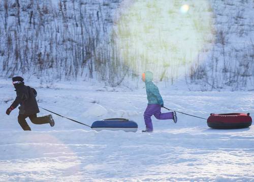 Two children run on snow while pulling colorful snow tubes behind them. Sunlight creates a lens flare effect.