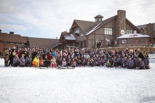 A large group of people in winter clothing poses on snow in front of a lodge, with skis and snowboards.