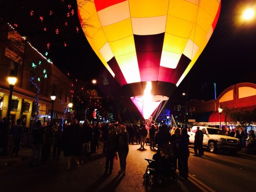 A colorful hot air balloon is illuminated at night, surrounded by a crowd and festive lights on a busy street.