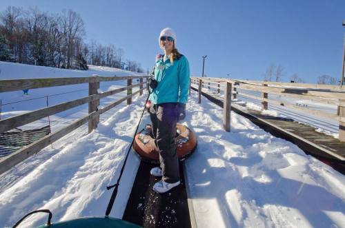 A woman stands on a snowy slope, preparing to go tubing, with a sunny blue sky in the background.
