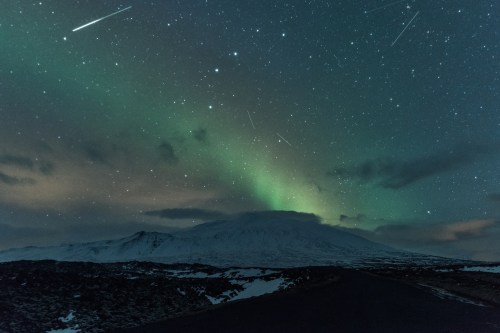 A snowy landscape under a starry sky, featuring the Northern Lights and shooting stars above a mountain.