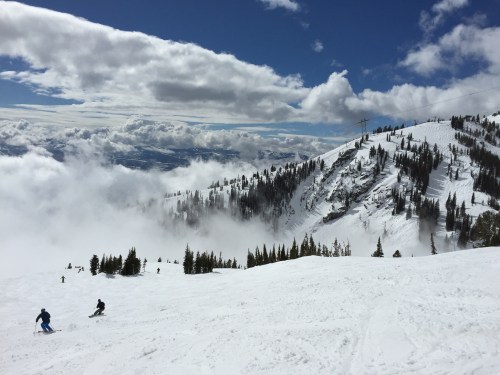 Skiers navigate a snowy slope with trees and clouds in the background under a bright blue sky.