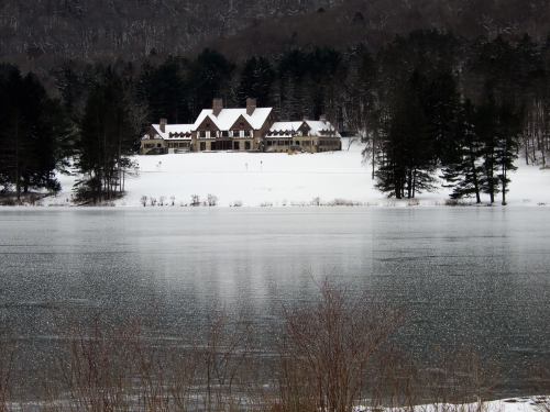 A large, snow-covered house sits by a frozen lake, surrounded by trees and a winter landscape.