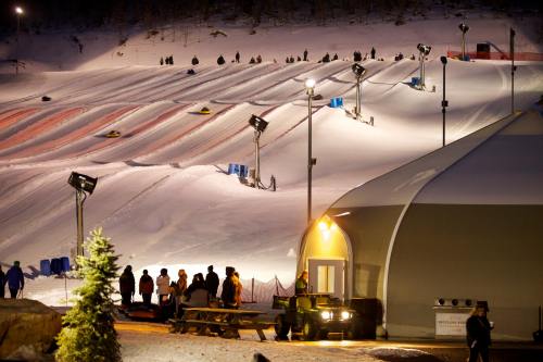 Snow tubing hill at night, with people sliding down and a warm building nearby, illuminated by bright lights.