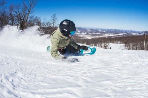 A snowboarder carves through fresh snow on a sunny day, with a scenic mountain backdrop.