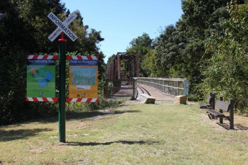 Signpost with trail information beside a wooden bridge, surrounded by greenery and a bench in a park setting.