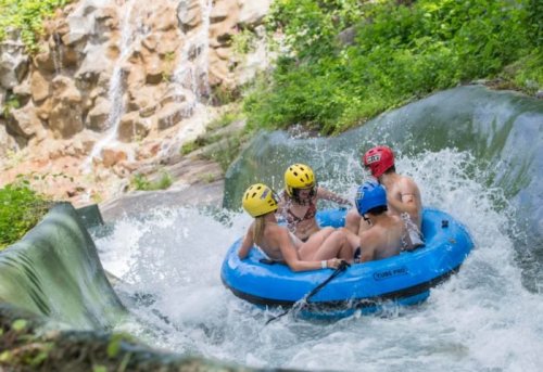 Four people in a blue inflatable raft navigate a water slide surrounded by greenery and a waterfall.