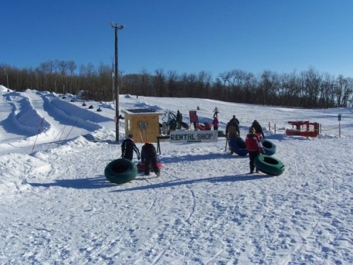 A snowy landscape with a rental shop sign, people carrying inner tubes, and sledding hills in the background.