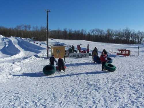 A snowy landscape with a rental shop sign, people carrying inner tubes, and sledding hills in the background.